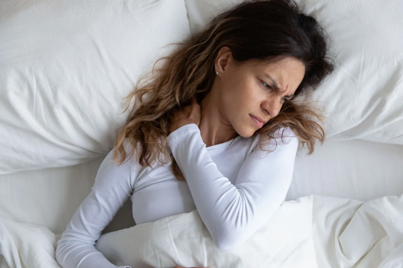 Woman in bed holding her neck in pain, illustrating discomfort from sleeping positions causing neck pain.